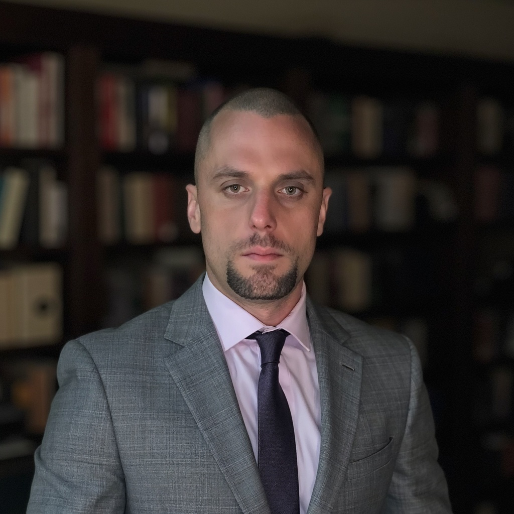 Michael wearing a grey suit and maroon tie infront of a book shelf